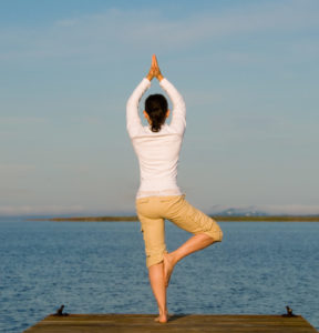 Frau in Yoga-Haltung auf einem Bein mit den Händen über dem Kopf gefaltet vor einem See stehend