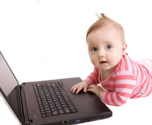 ein auf dem Bauch liegendes Baby hat eine Hand auf dem Laptop, der vor ihm am Boden aufgeklappt steht, schaut in die Kamera