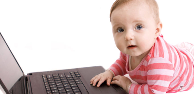 ein auf dem Bauch liegendes Baby hat eine Hand auf dem Laptop, der vor ihm am Boden aufgeklappt steht, schaut in die Kamera