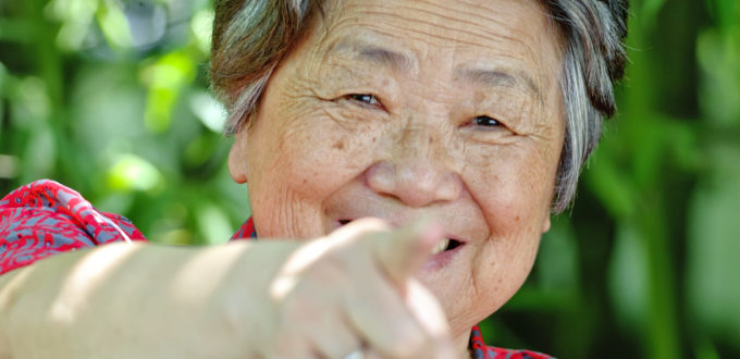 eine alte Frau zeigt mit großem Lachen mit der gestreckten Hand und dem Zeigefinger voll in die Kamera
