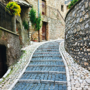 eine mit blauen Ziegelsteinen gepflasterter Gehweg führt nach oben, rechts einer Steinmauer entlang, links hängen Blumen in den altbürgerlichen Häusern, mutet nach einem Inselstädtchen an