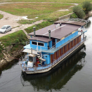 ein hellblaues Hausboot mit zwei Rauchfängen liegt am Ufer eines Flusses. Solarpaneelen auf dem Dach, eine Wendeltreppe außen ins obere Stockwerk. Man sieht auch einen Griller an Deck.