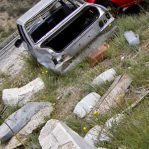 ein ehemals weißes Schrottauto steht auf einem mit Gras und sogar einzeln mit gelbem Löwenzahn überwachsenen Schottergrund.