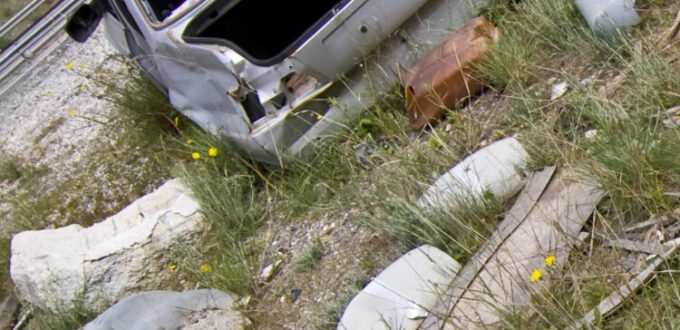 ein ehemals weißes Schrottauto steht auf einem mit Gras und sogar einzeln mit gelbem Löwenzahn überwachsenen Schottergrund.